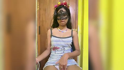 Media: Video of a young woman with medium brown skin, wearing a black lace mask, floral headband, and a striped dress, sitting in a wooden chair.