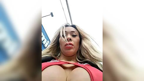 Media: A low-angle video of a blonde woman with large breasts wearing a red top, taken outdoors against a bright sky.