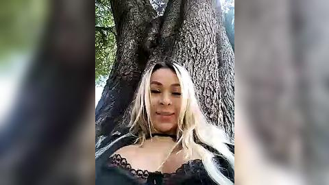 Media: Video of a blonde woman with medium-length hair, wearing a black lace bralette, leaning against a large, textured tree trunk in a forest, smiling.