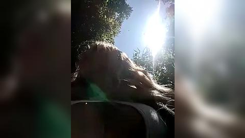 Media: A blurry video of a person with long, blonde hair, wearing a white shirt, captured outdoors in a bright, sunny environment with sun flares and green foliage in the background.