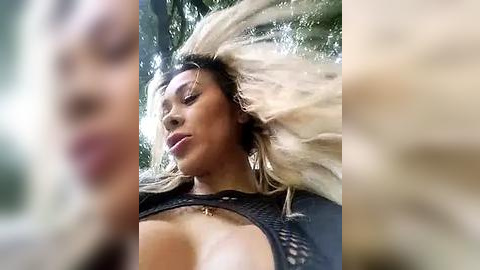 Media: A video of a woman with long, blonde hair, tanned skin, and large breasts, wearing a black, fishnet top, captured in a blurred, outdoor setting with a focus on her dynamic, wind-blown hair.