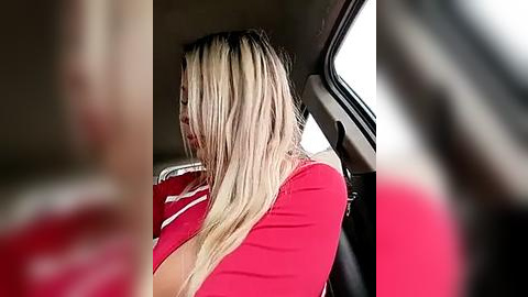 Media: A video of a woman with long, blonde hair, wearing a red shirt, sitting in a car. The background is blurry.