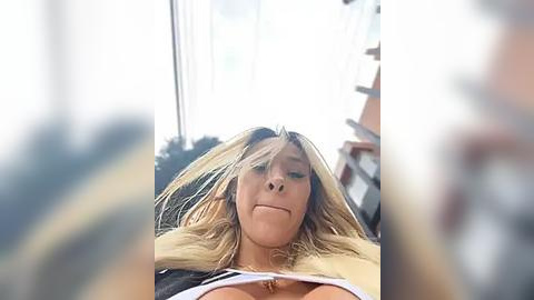 Media: Video of a blonde woman lying on her back, wearing a white top, with her hair covering her face, taken from a low angle, blurred background.