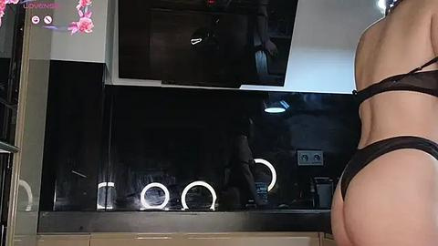 Media: Video of a woman with a toned, pale skin tone wearing black lingerie, standing with her back to the camera in a modern kitchen with black appliances, illuminated by ring lights.