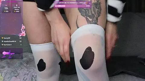 Media: Video of a person with light skin, wearing white thigh-high stockings with black cow print, holding a tattooed leg. Background includes a dark couch and a purple screen with social media icons.