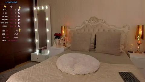 Media: Video of a luxurious, dimly lit bedroom with a plush, white tufted headboard, beige bedding, and gold lamps on nightstands. A large, mirrored vanity with glowing bulbs is on the left. A furry white pillow is centered on the bed.