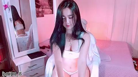 Media: Video of a young Asian woman with long black hair, wearing a white crop top and panties, sitting on a bed in a softly lit bedroom.