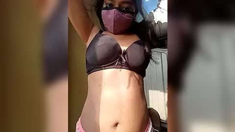 Media: Video of a woman with medium brown skin and curly hair wearing a black bra, pink panties, and a purple face mask outdoors. Background includes a beige wall and a gray structure.