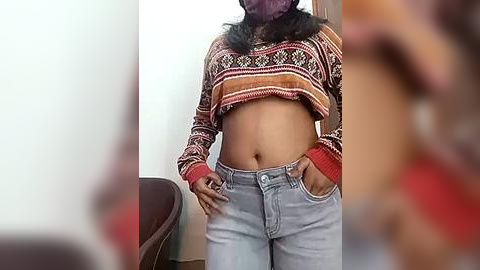 Media: A video of a young woman with medium brown skin, wearing a patterned, cropped sweater and light blue jeans, standing in a room with a blurred background.