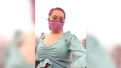 Media: Video of a young woman with light brown skin, wearing a purple face mask and a light green blouse with ruffled sleeves. Background features blurred urban buildings.