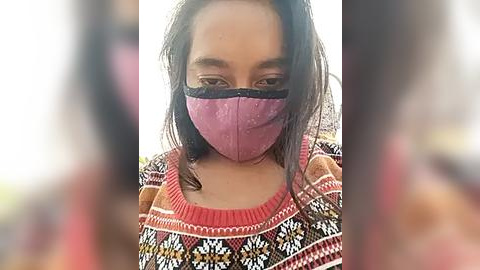 Media: Video of a young woman with long dark hair, wearing a pink face mask and a colorful patterned sweater, against a blurry background.