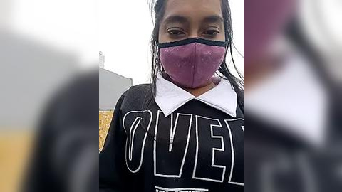 Media: Video of a young woman with dark skin, wearing a black hoodie with \"Olive\" in white letters, a purple face mask, and a white collared shirt. Background is blurry.