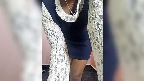 Media: Video of a person with medium brown skin wearing a navy blue dress with lace sleeves and a white lace collar, standing indoors against a pink wall.