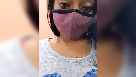 Media: Video of a young woman with medium brown skin, wearing a pink face mask and a gray sweater adorned with blue flowers, set against a blurred peach background.