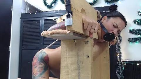 Media: Video of a nude woman with a colorful tattoo on her backside, restrained in a wooden X-frame, gagged with a ball, and wearing a blindfold, in a festive room with Christmas decorations.