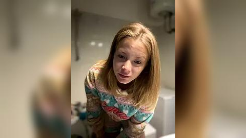 Media: Video of a young woman with long, straight blonde hair, wearing a colorful, patterned sweater, leaning forward with a curious expression. The background features a white-tiled bathroom with a sink and mirror.