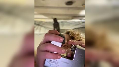 Media: Video of a woman taking a selfie with a white iPhone in an airplane bathroom, holding the phone with both hands. The background is blurred, showing the airplane's interior.