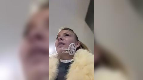 Media: Video of a woman with blonde hair wearing a yellow faux fur coat and a large, ornate silver earring shaped like a butterfly, gazing upward.