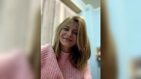 Media: Video of a smiling Caucasian woman with long blonde hair, wearing a pink knitted sweater, standing in a softly lit room with blue walls and a curtain.