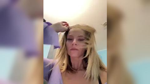 Media: Video of a blonde woman with fair skin, wearing a light purple shirt, brushing her hair in a bathroom with blue tiles.