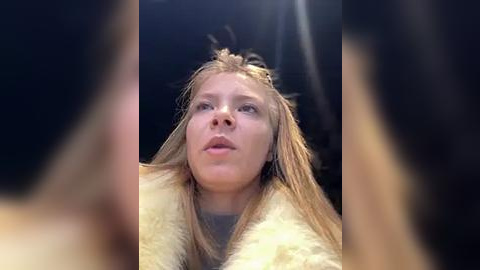 Media: Video of a blonde woman with straight hair, wearing a fur coat, looking up with a neutral expression, set against a dark background.