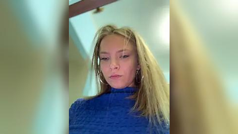 Media: A video of a young woman with long blonde hair, wearing a blue turtleneck sweater, standing in a blurred hallway, looking serious.