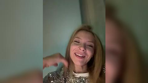 Media: Video of a young woman with straight, shoulder-length blonde hair, wearing a sparkly silver sequin top, smiling widely with her hands up, in a dimly lit room with a teal wall.