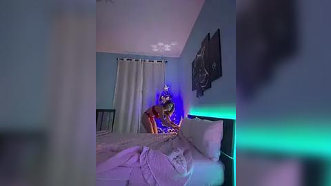 Media: A video captures a dimly lit bedroom with a woman in a red dress and black top, bent over a bed, illuminated by blue light. The room features a black headboard, white curtains, and a wall-mounted TV.