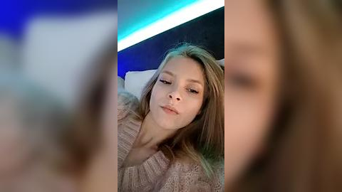 Media: Video of a young Caucasian woman with straight, shoulder-length blonde hair, wearing a cozy, light-colored knitted sweater, reclining on a bed with a dark headboard. The background features a modern, blue and white light strip.