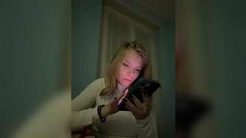 Media: A video of a young blonde woman with fair skin, wearing a beige sweater, intently using a smartphone in a dimly lit room with teal walls and a white curtain in the background.