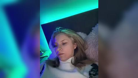 Media: Video of a young, fair-skinned girl with long blonde hair, wearing a white turtleneck, lying on a dark bed with a fluffy pillow, under blue and green lighting.