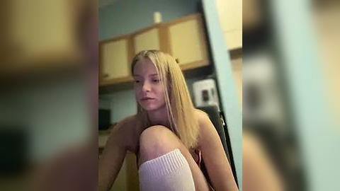 Media: Video of a blonde woman in a kitchen, sitting topless with her legs crossed, wearing white knee-high socks. Background shows wooden cabinets and a microwave.