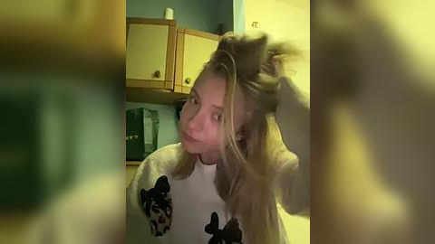 Media: A video of a blonde woman in a white sweater with a black cat design, holding her hair, in a dimly lit kitchen with wooden cabinets and a green coffee maker.