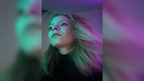 Media: A video of a woman with messy, blonde hair, wearing a black top, standing in a dimly lit room with green and purple lighting, creating a surreal, ethereal atmosphere.