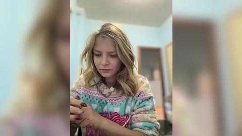 Media: A video of a young blonde girl in a colorful, patterned sweater, sitting on a bed in a softly lit room with pastel walls and a wooden door in the background.