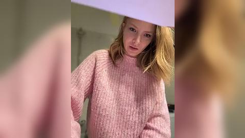 Media: Video of a young, fair-skinned, blonde-haired girl in a pink, textured sweater, looking solemnly into a mirror, with out-of-focus blurred figures in the background.