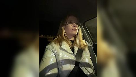 Media: Video of a blonde woman in a shiny white puffer jacket, driving at night. Her expression is contemplative, and the car's interior is dimly lit.