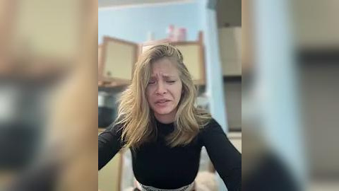 Media: A video shows a woman with shoulder-length blonde hair, wearing a black top, looking distressed in a blurred kitchen setting with cabinets and a stove in the background.