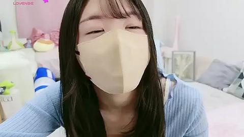 Media: A video of an East Asian woman with long black hair and fair skin, wearing a beige surgical mask indoors. She is in a soft, light blue sweater, in a cozy, slightly cluttered room with pastel decor.