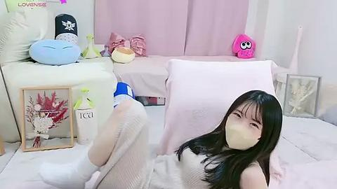 Media: Video of a young Asian woman with long black hair, wearing a beige knitted dress and white socks, lying on a bed with pink sheets, surrounded by plush toys and a framed art piece.