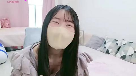 Media: A video of an East Asian woman with long black hair, wearing a beige mask and a gray sweater, sitting on a bed in a cozy room with pastel-colored pillows.