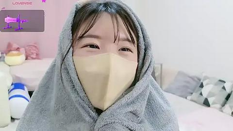 Media: Video of an Asian woman with short black hair, wearing a beige surgical mask, wrapped in a grey towel, in a softly lit bedroom with white walls, pink pillows, and a bed.