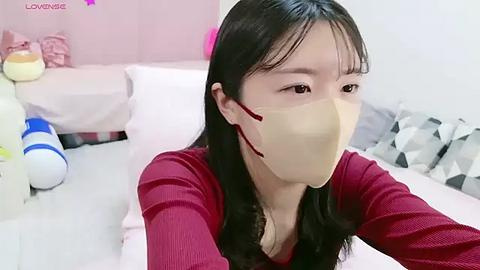 Media: Video of an East Asian woman with long black hair, wearing a beige face mask and a red long-sleeved shirt, in a pink-themed bedroom with toys and geometric-patterned pillows.