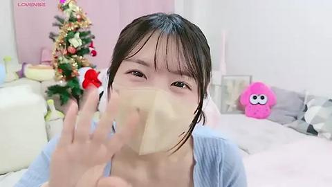 Media: Video of a young East Asian woman with light skin, brown hair, and bangs, wearing a face mask, sitting in a cozy, festive room with a decorated Christmas tree, plush toys, and a pink plushie in the background.