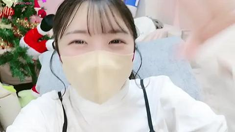 Media: A video of an East Asian woman with dark hair, wearing a beige surgical mask and a white lab coat, standing in a brightly lit room with Christmas decorations and a fluffy white carpet.