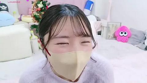 Media: Video of an Asian woman with black hair and bangs, wearing a yellow face mask, lying on a bed with a white blanket, surrounded by pink and white plush toys.