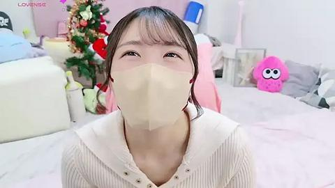 Media: Video of an East Asian woman with short, dark hair wearing a beige face mask and white ribbed sweater, sitting on a bed in a pink-themed room with a Christmas tree and plush toys.