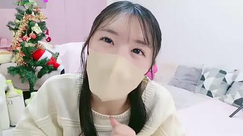 Media: A video of an Asian woman with long black hair, wearing a beige face mask, sitting on a bed in a cozy room with a decorated Christmas tree and white pillows.