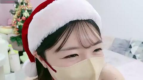 Media: Video of an Asian woman with fair skin, dark hair, wearing a red Santa hat, and a beige surgical mask, in a bright, festive room with a Christmas tree and white couch.