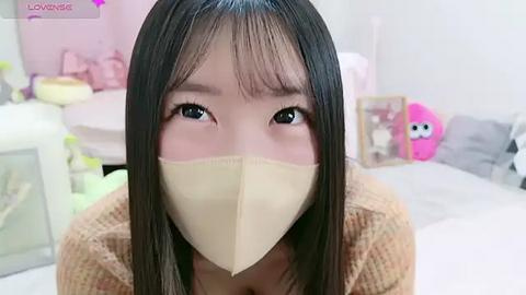 Media: A video of an East Asian woman with long black hair, wearing a beige mask, in a pastel-themed bedroom with plush toys.